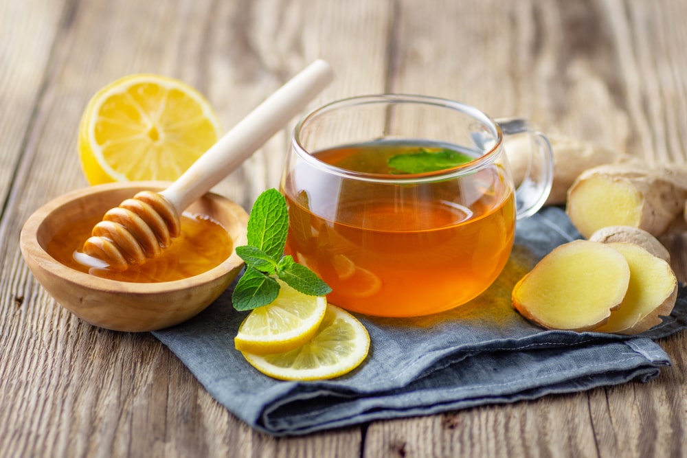 A glass cup of tea with lemon, mint, ginger and honey on top of a blue cloth on a wooden table.