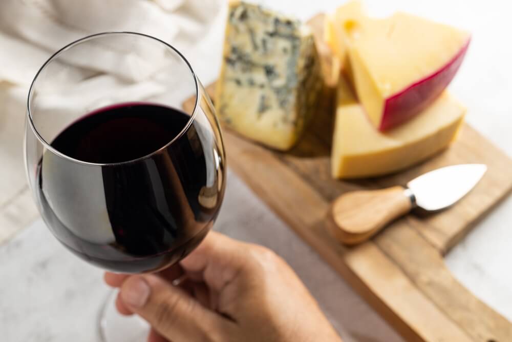 an image of a woman holding a glass of red wine with a board of 3 cheese varieties blurred in the back