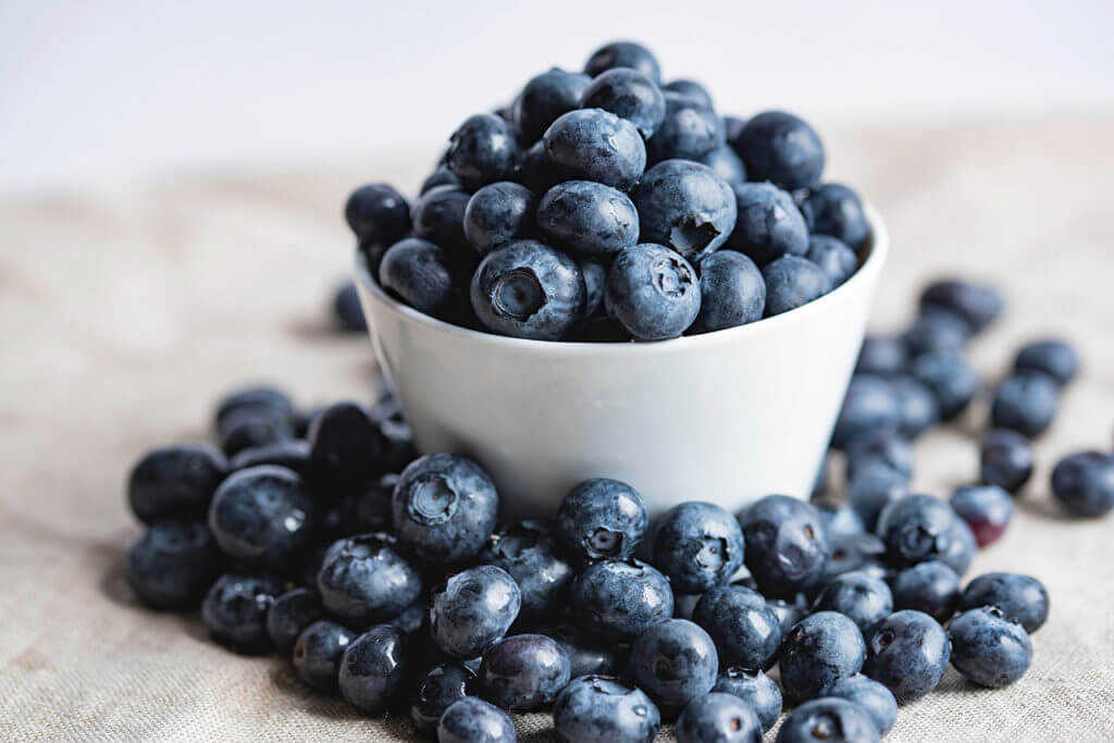 A small white bowl with blueberries overflowing onto cloth mat.