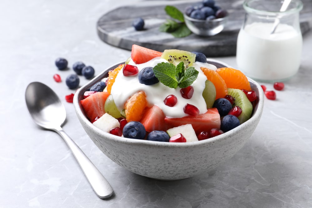 A bowl of fruit with white yogurt on top. The bowl is on a table between a spoon and a glass of yogurt.