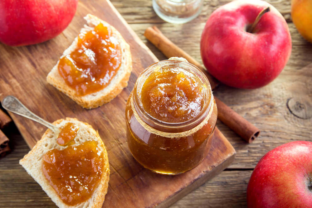 jar of amish apple butter on a cutting board next to a slice of bread with apple butter spread on top