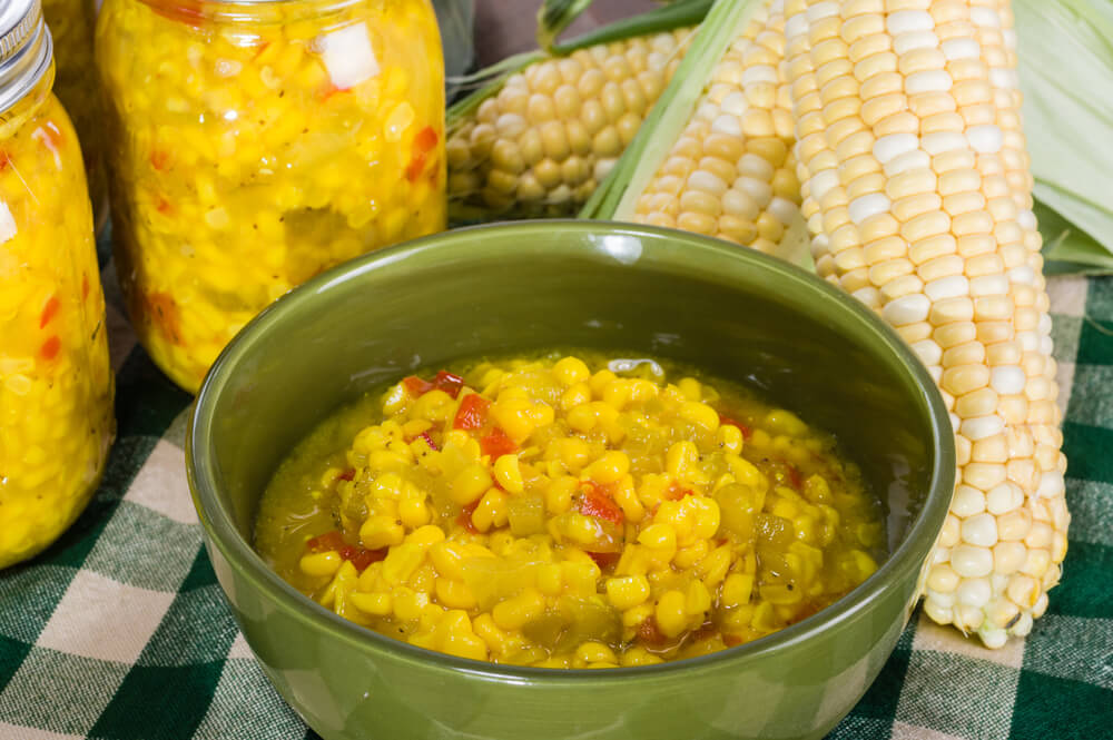 a bowl of sweet corn relish resting next to a large jar of relish and some corn on the cob
