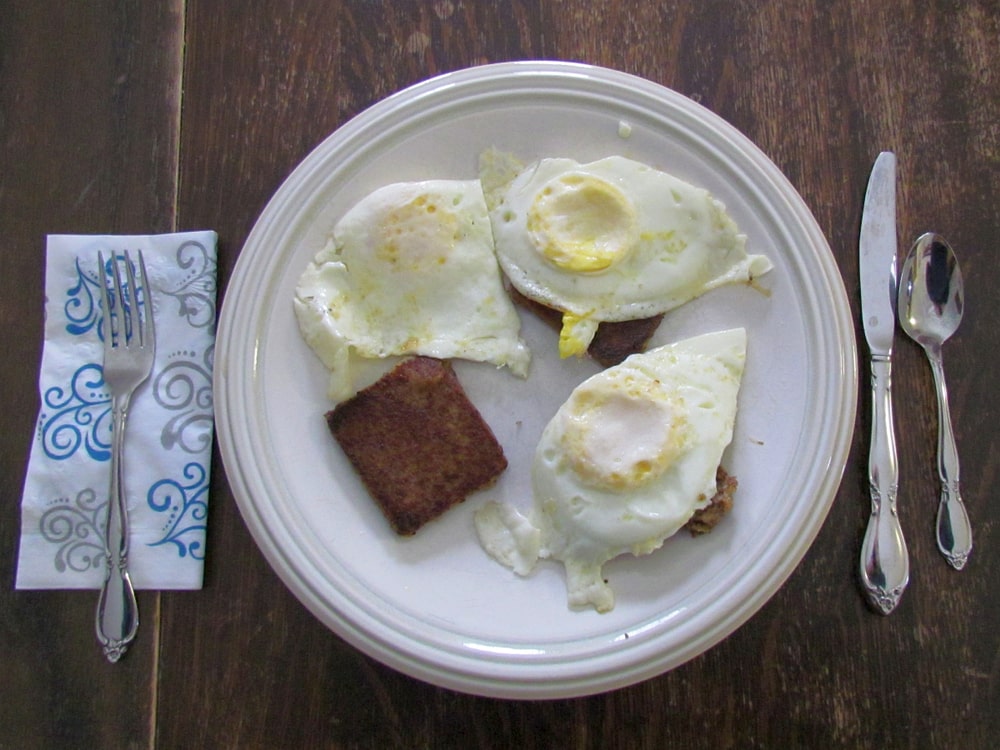 Scrapple and over-easy eggs on a white plate with silverware and napkin